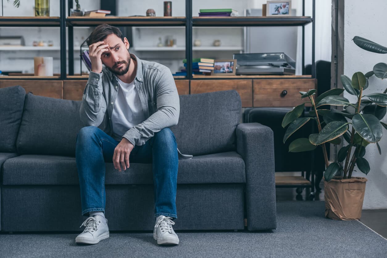 depressed man sitting on couch, touching face and grieving at home